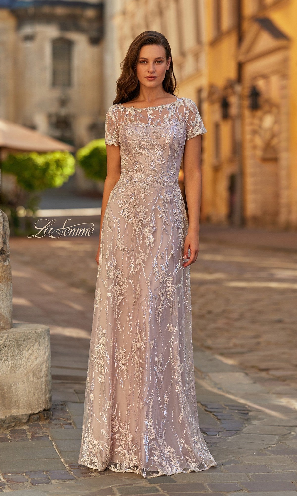 A woman stands on a cobblestone street wearing the La Femme 31838 long formal dress in light mauve, featuring short sleeves, an A-line silhouette, and intricate beaded embroidery with silver detailing.